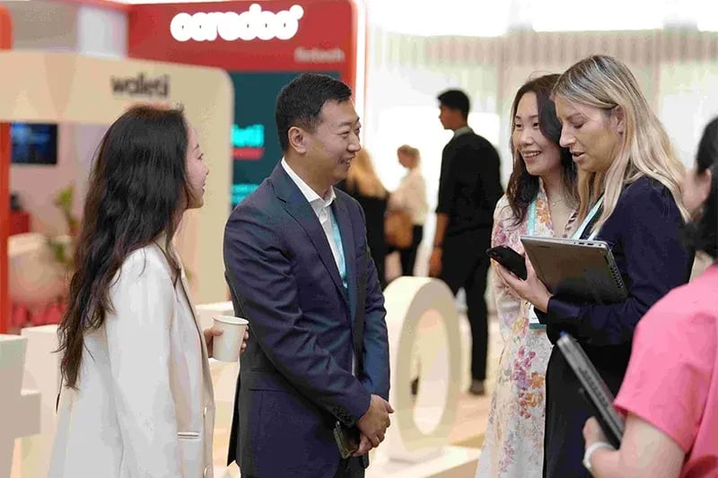A group of professionals engaged in conversation at a networking event, with a man in a suit smiling and talking to three women—one holding a coffee cup and another holding a tablet. The background shows a busy exhibition area with branding from Ooredoo.