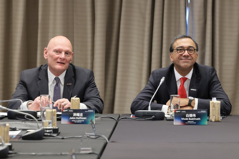 John Hoffman and Vivek Badrinath seated at a conference table during the MWC25 Doha Digital Leaders Programme, both smiling and facing the camera. Nameplates in English and Chinese are visible in front of them.
