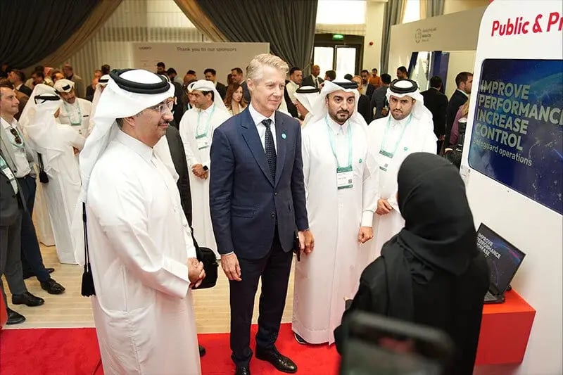 A group of dignitaries and attendees visit a technology exhibition booth at a conference. A man in a blue suit stands alongside officials in traditional white Middle Eastern attire, attentively listening to a woman in a black abaya who is presenting information. A display screen at the booth reads 'IMPROVE PERFORMANCE, INCREASE CONTROL, Safeguard operations.' The scene is professional and formal, indicating a high-level industry event, likely part of the M360 MENA series.