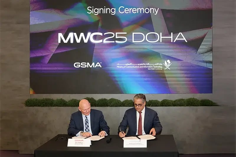 Two men seated at a table signing documents during the MWC25 Doha signing ceremony. A large digital screen behind them displays the event name 'MWC25 DOHA' along with logos of GSMA and the Ministry of Communications and Information Technology. The man on the left wears a dark suit and striped tie, while the man on the right wears glasses, a dark suit, and a red tie. Both are focused on signing the documents.