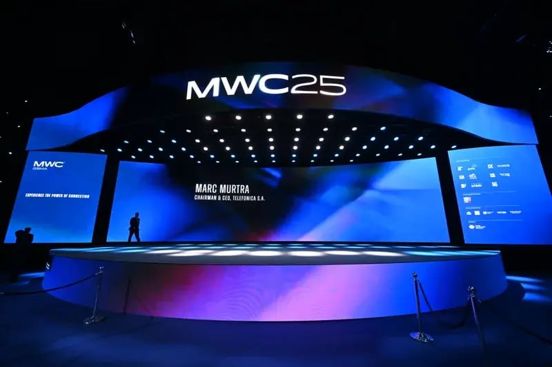 Large stage setup at MWC25 with immersive blue lighting and digital screens displaying the name Marc Murtra, Chairman & CEO of Telefónica S.A., during a keynote session.