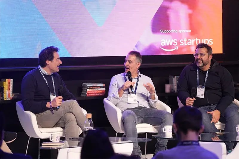 Three men speaking on stage during a startup panel supported by AWS Startups, each holding microphones and seated in front of a large screen.