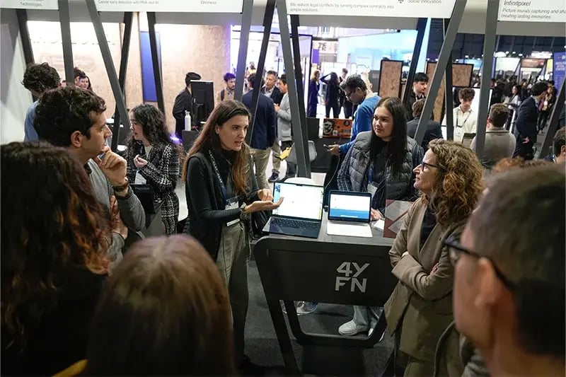 Woman presenting at a 4YFN booth to a small crowd, with open laptops displaying content and branding above the booth.