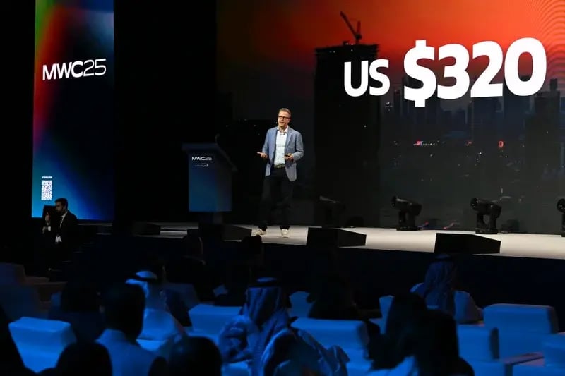 Man in a blue blazer presents on stage at MWC25. Large screen displays "US $320." Audience watches attentively, room dimly lit, tech-centric atmosphere.