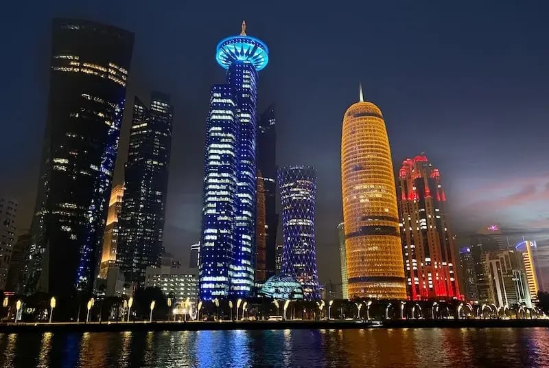 Night view of the West Bay skyline in Doha, Qatar, showcasing illuminated skyscrapers in vibrant blue, gold, and red hues reflecting off the waterfront.