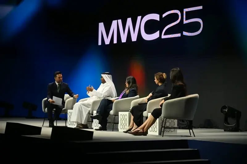 Panelists seated on stage at MWC25 conference, engaged in discussion. Large "MWC25" text displayed above, with a diverse, attentive audience.