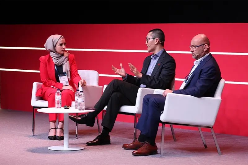 Three panelists engage in a discussion on stage at a conference with a red backdrop. A woman in a red suit and beige headscarf listens attentively while two men, dressed in business attire, speak and gesture. Bottled water and microphones are placed on a small table in front of them.