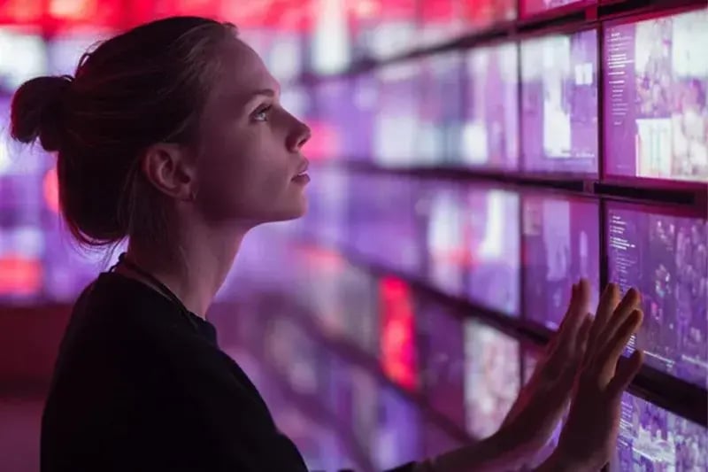 Woman touching a wall of interactive digital screens with red and purple lighting, symbolizing data and future technology.