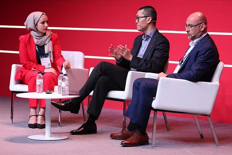 Three speakers in discussion on stage during MWC, seated on white chairs against a red backdrop, with bottled water on a round table.