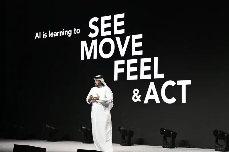 Speaker presenting on stage at MWC Doha with a large screen displaying the message “AI is learning to see, move, feel & act” in bold text behind him.