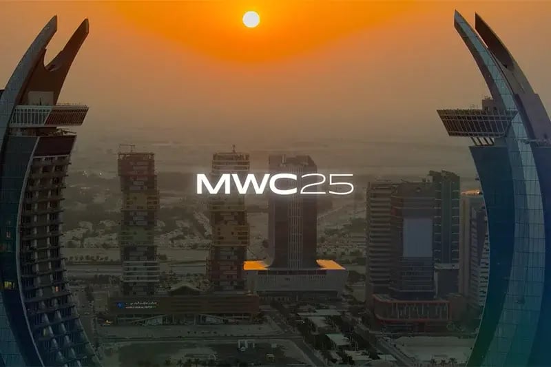 Sunset view over a modern city skyline with two curved skyscrapers in the foreground and several high-rise buildings in the background, with 'MWC25' text prominently displayed in the center.