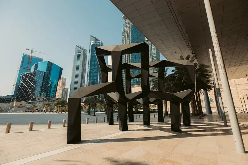 Modern outdoor sculpture with geometric black structures stands near tall, glass skyscrapers under a clear blue sky, conveying a sleek, urban vibe.