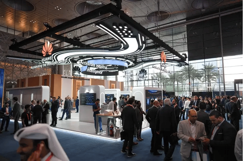 Large tech booth at a convention, featuring futuristic lighting and Huawei branding. Crowds of people engage and converse, showcasing a lively atmosphere.
